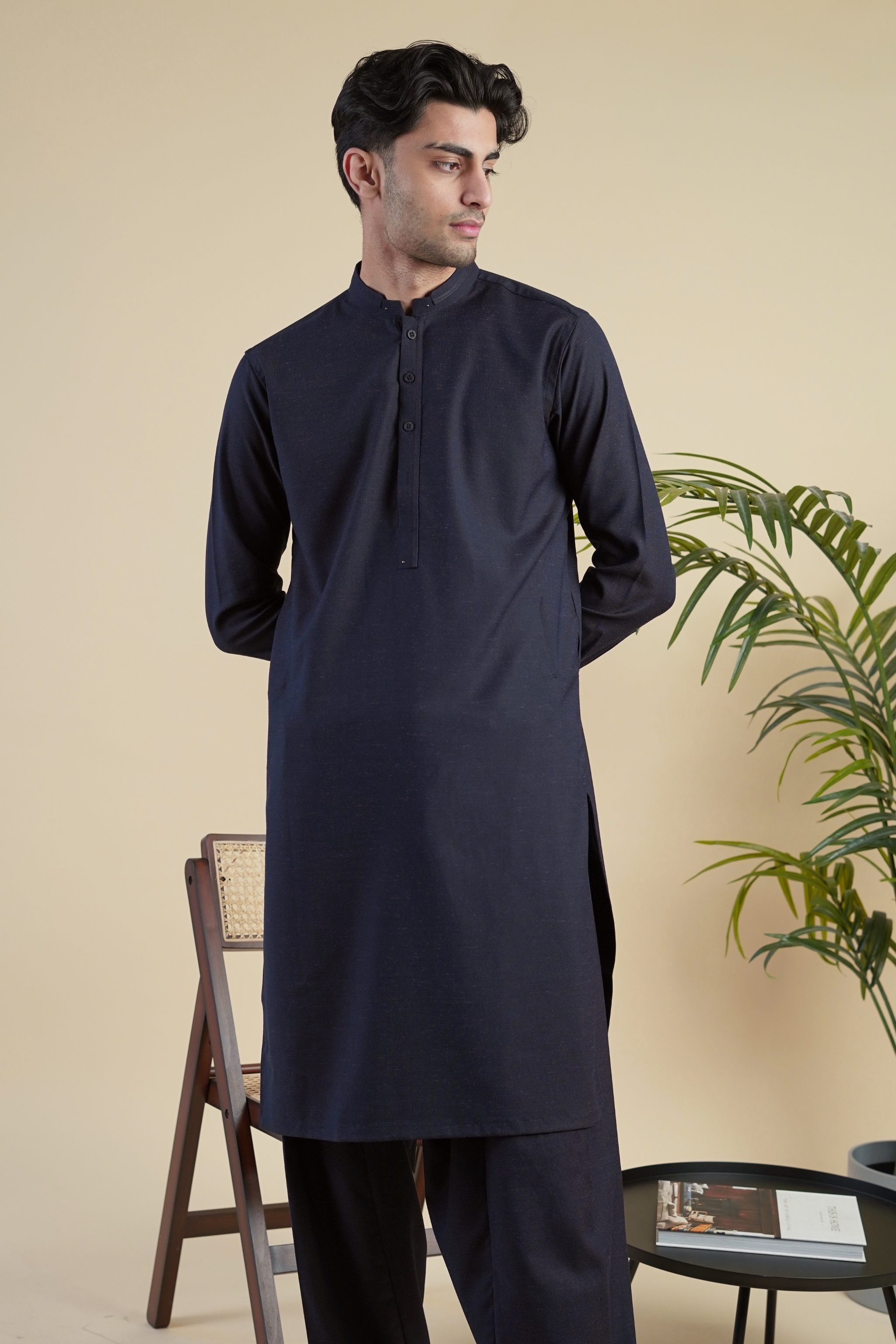 Pakistani male model standing wearing UD Fabrics men's navy blue shalwar kameez with purple undertones . Shalwar kameez with design on collar. Model poses looking away from camera. Men's pakistani clothing. Navy with purple undertones shalwar kameez. Design on collar of shalwar kameez.