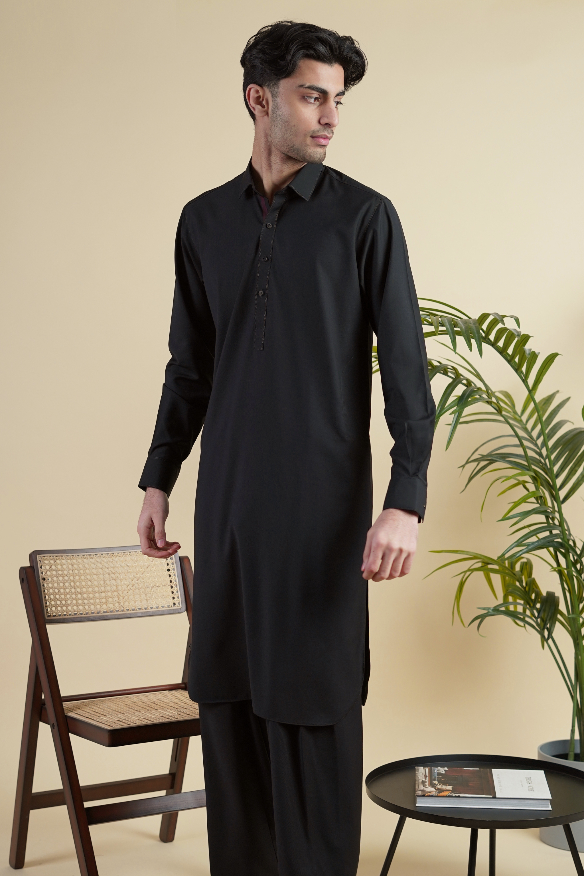 Pakistani male model standing wearing UD Fabrics men's black shalwar kameez. Shalwar kameez with full collar. Model poses looking away from camera. Men's pakistani clothing. Black shalwar kameez. Full collar shalwar kameez.