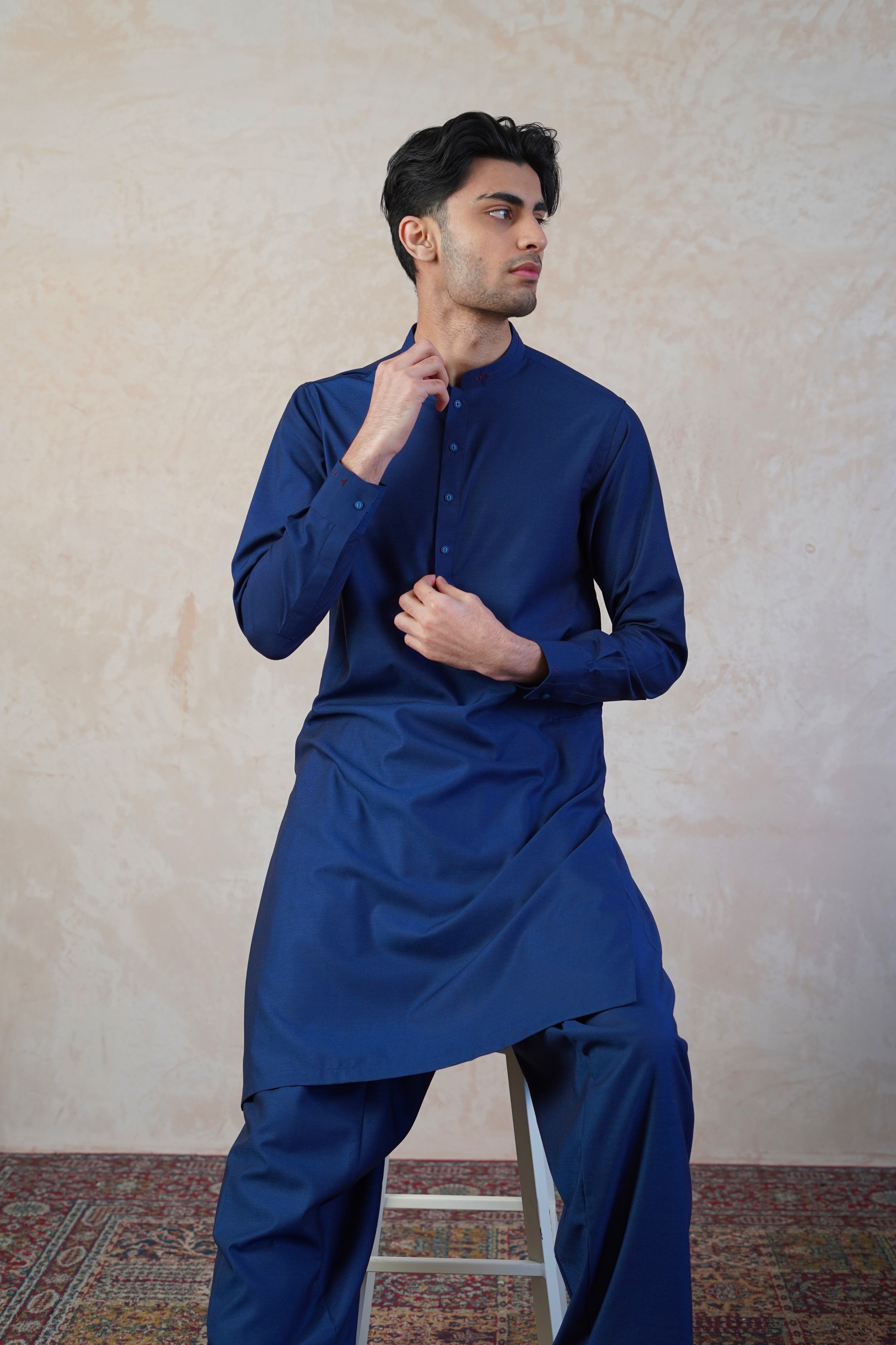 Pakistani male model sat on stool wearing UD Fabrics men's blue shalwar kameez. Shalwar kameez with design on collar. Model poses looking towards camera. Pakistani men's clothing
