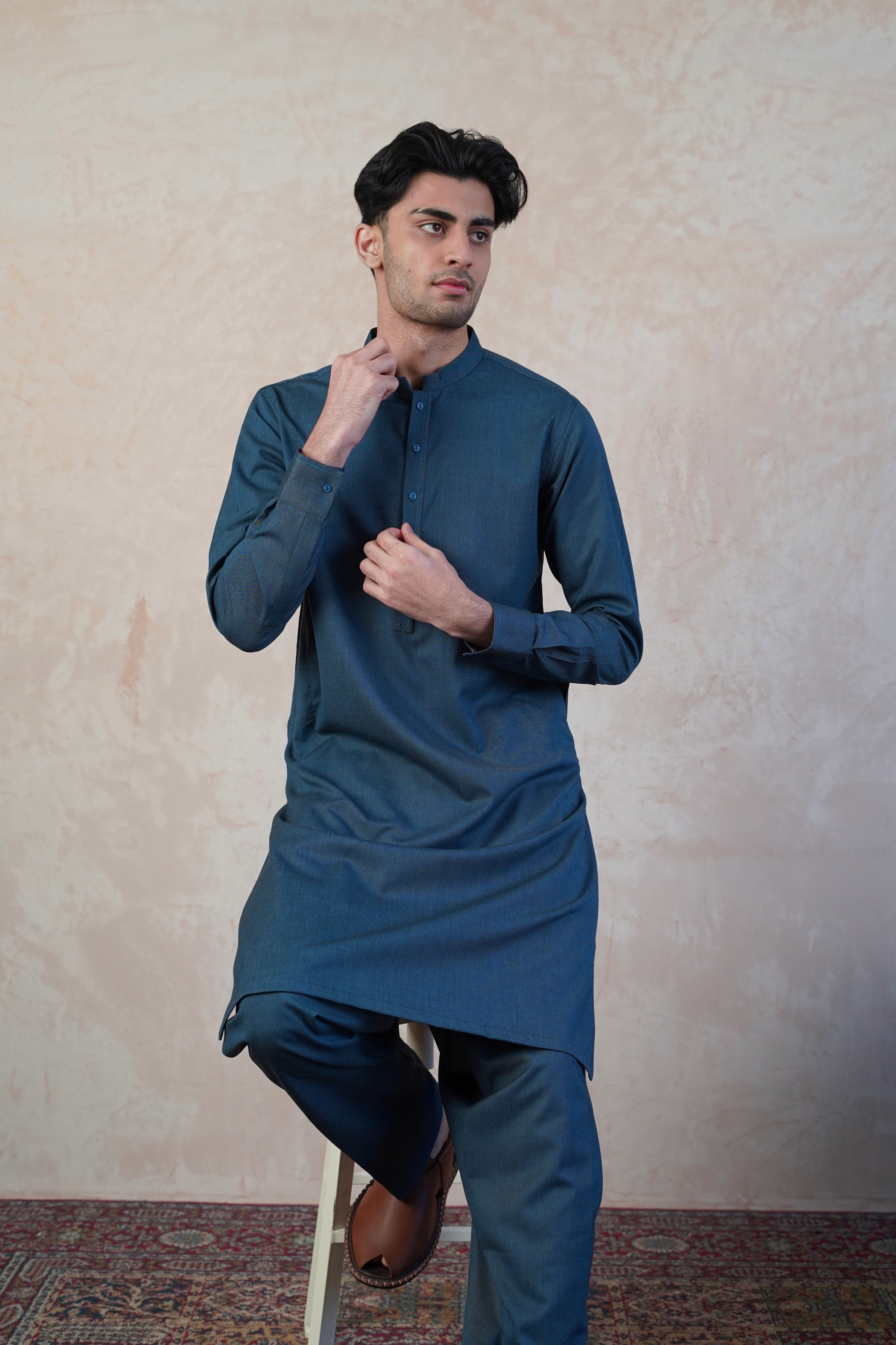 Pakistani male model sat on stool wearing UD Fabrics men's blue shalwar kameez. Shalwar kameez with design on collar. Model poses looking away from camera. Men's pakistani clothing.