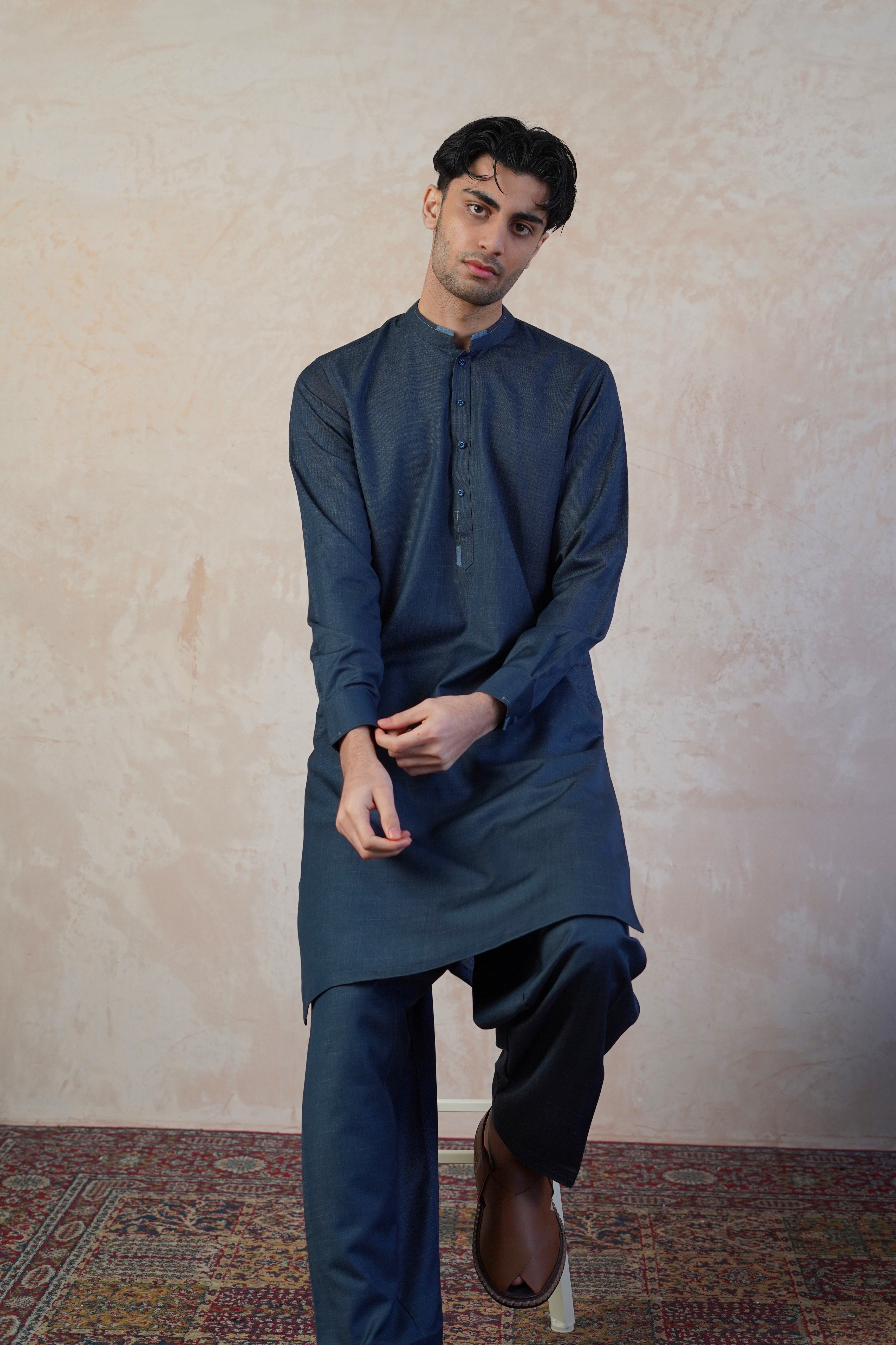 Pakistani male model sat on stool wearing a UD Fabrics deep aqua blue shalwar kameez with lighter blue design on the collar