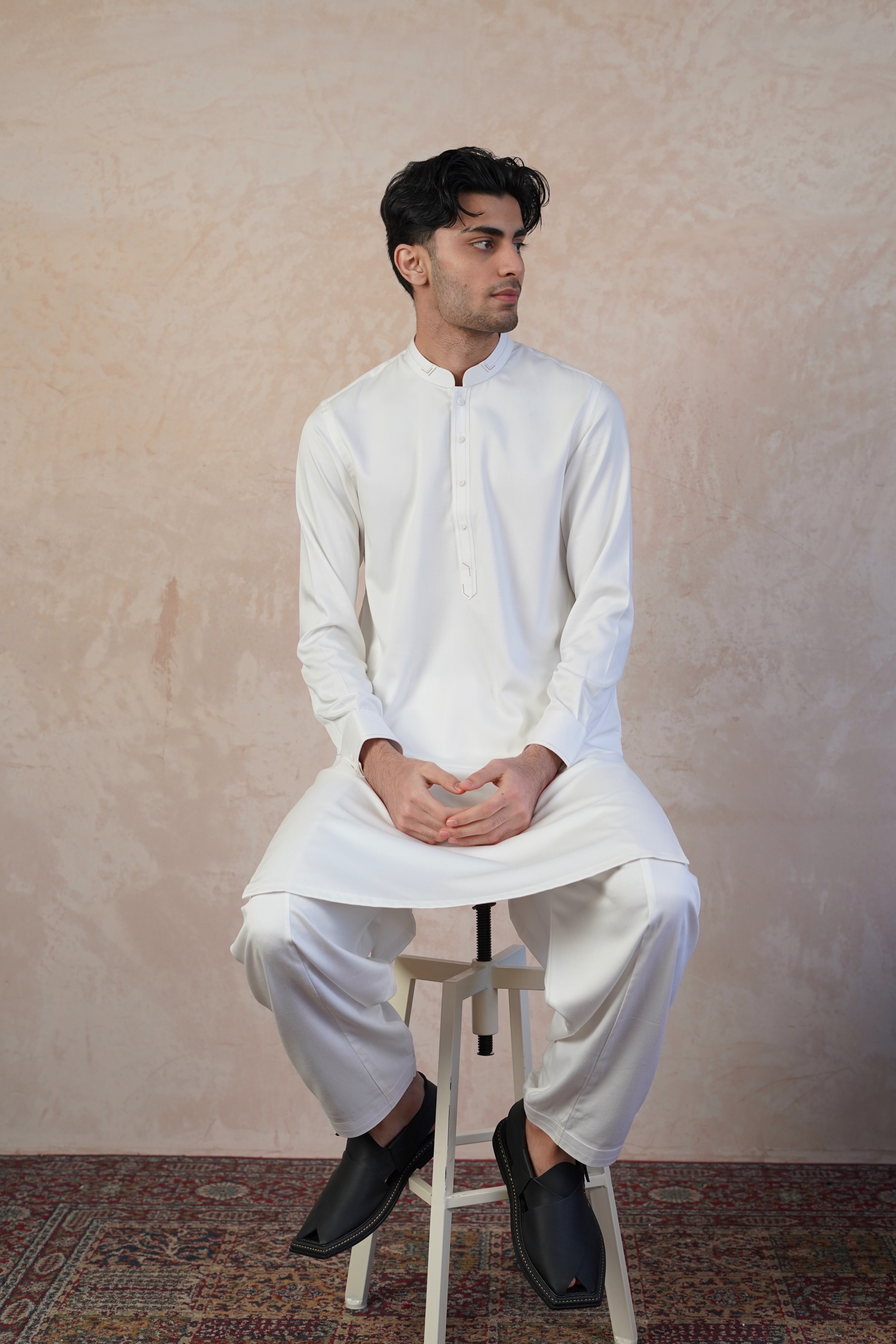 Pakistani male model sitting on stool wearing UD Fabrics men's white shalwar kameez. Shalwar kameez with design on collar. Model poses looking away from camera. Men's pakistani clothing. White shalwar kameez