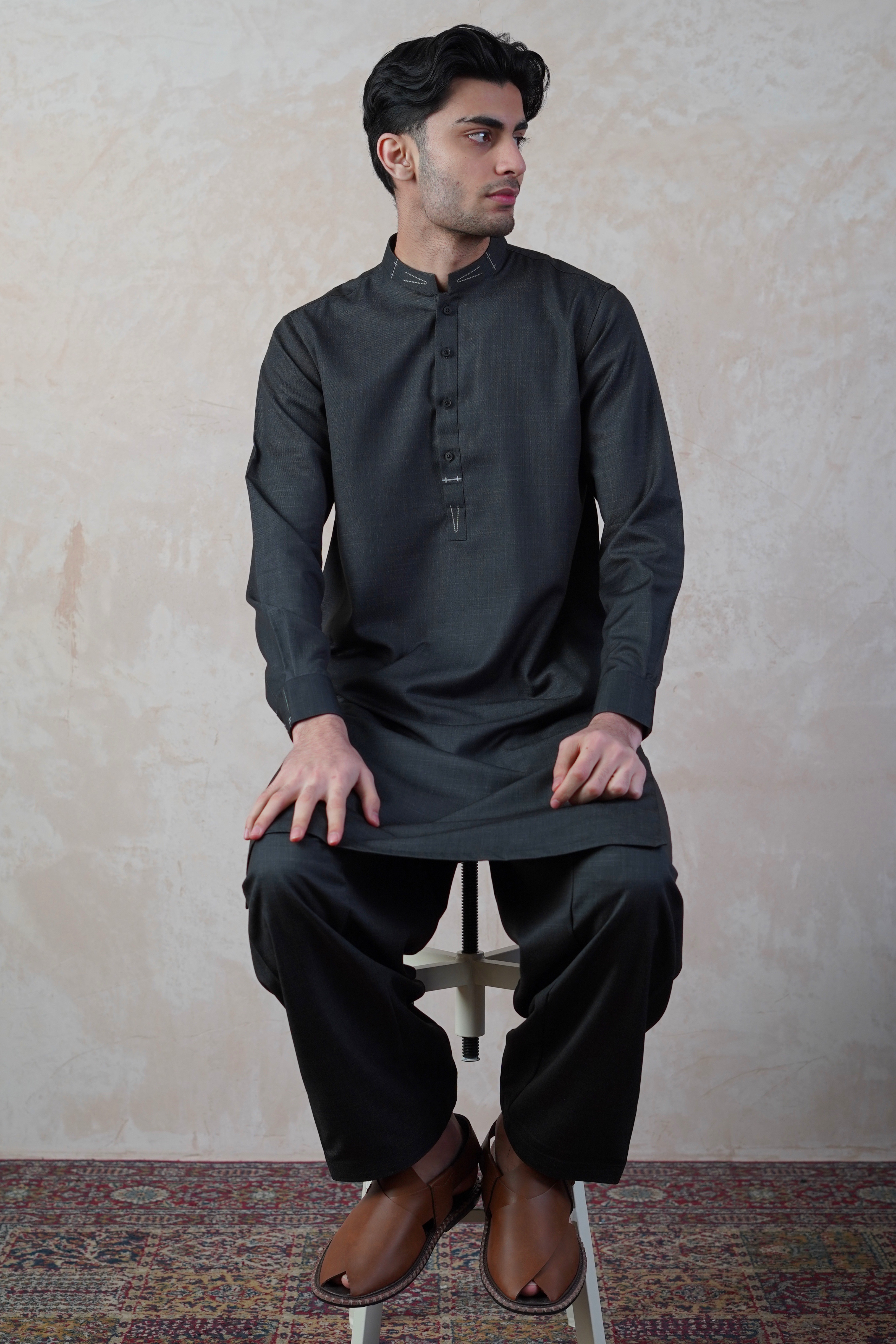 Pakistani male model sat on stool wearing UD Fabrics men's black shalwar kameez. Shalwar kameez with design on collar. Model poses looking away from camera. Men's pakistani clothing. Black shalwar kameez