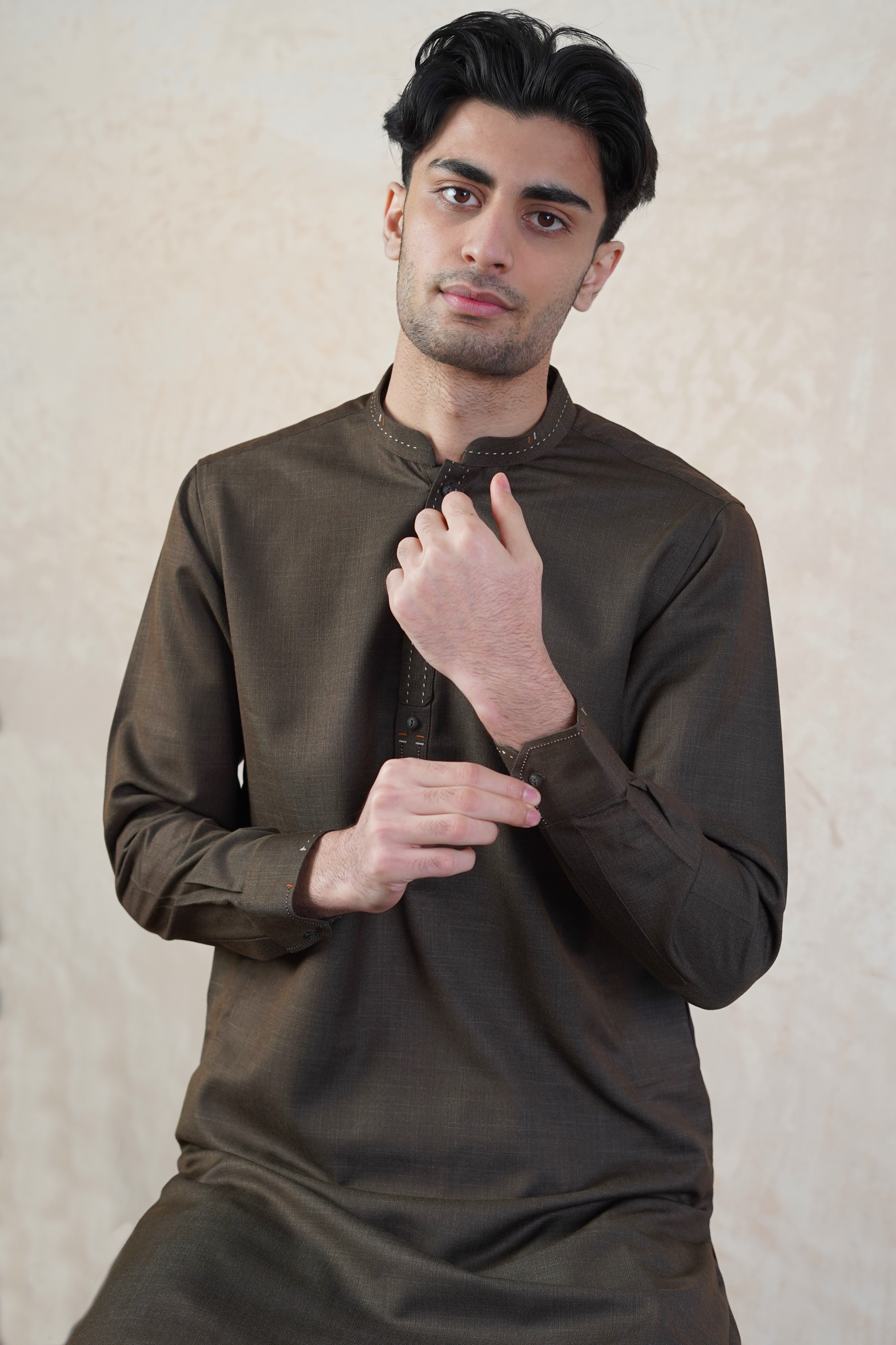 Pakistani male model standing wearing UD Fabrics men's brown shalwar kameez. Shalwar kameez with design on collar. Model poses looking towards camera. Men's pakistani clothing. Brown shalwar kameez. Design on collar of shalwar kameez.