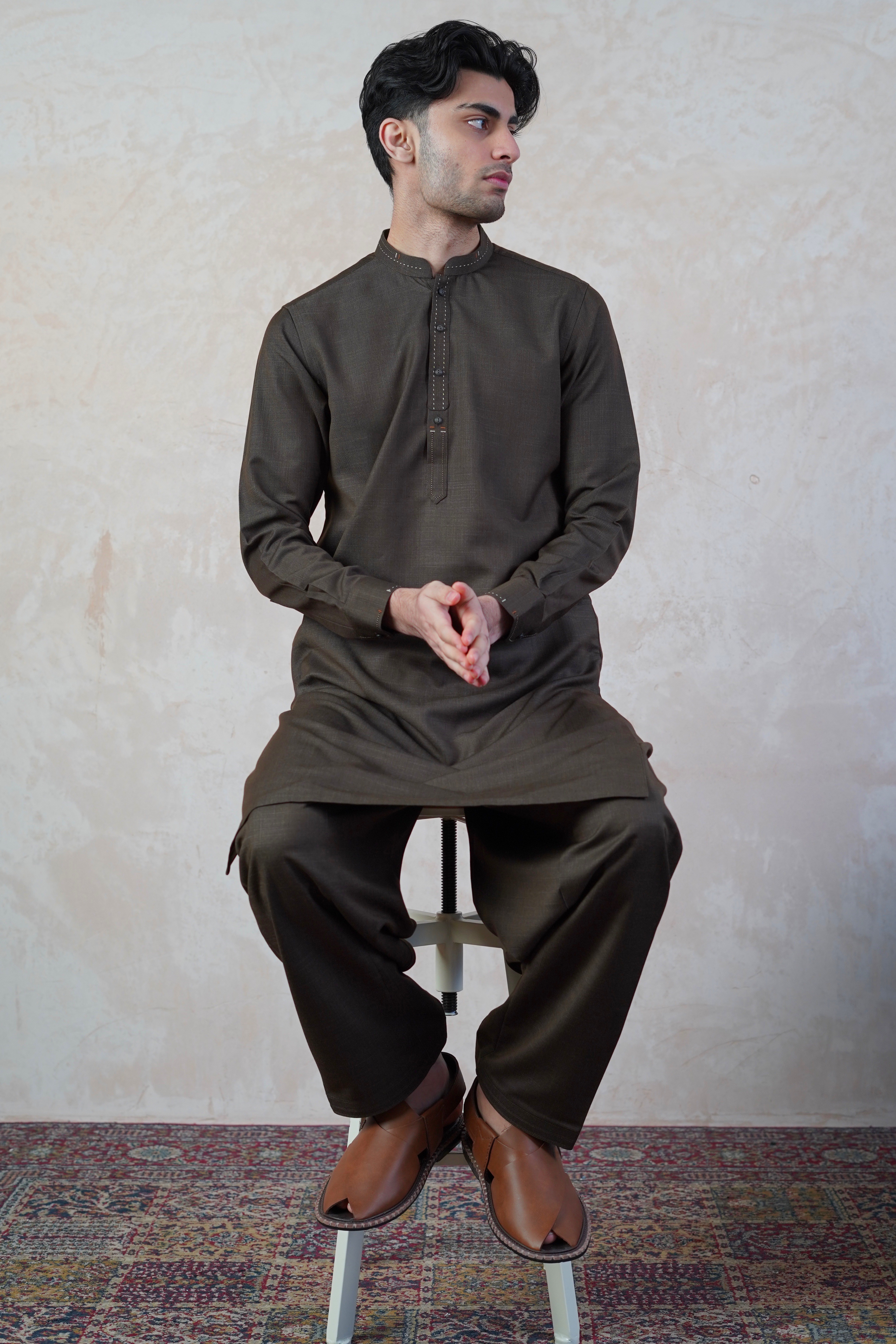 Pakistani male model standing wearing UD Fabrics men's brown shalwar kameez. Shalwar kameez with design on collar. Model poses looking away from camera. Men's pakistani clothing. Brown shalwar kameez. Design on collar of shalwar kameez.