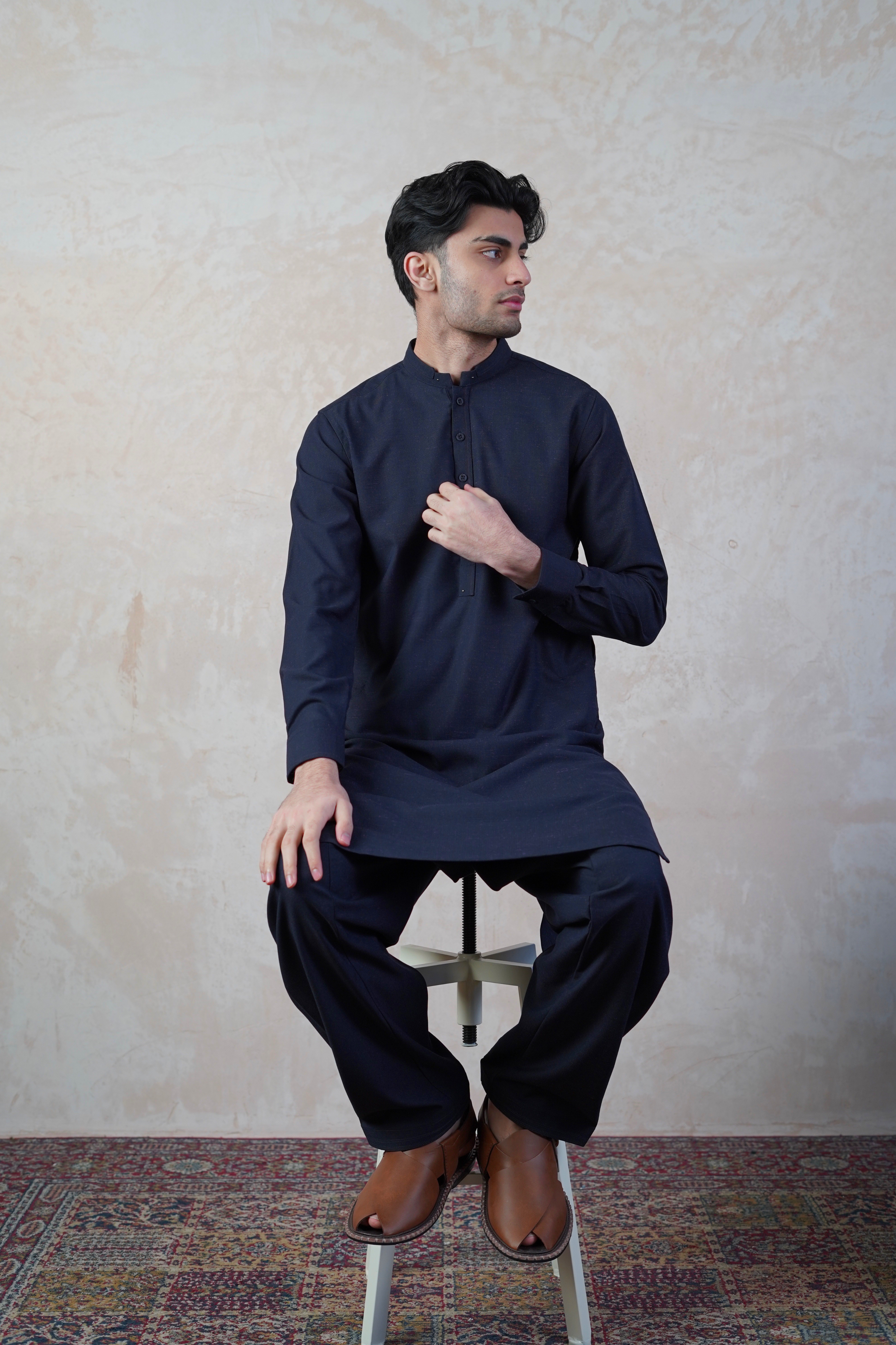 Pakistani male model sat on stool wearing UD Fabrics men's navy blue shalwar kameez with purple undertones . Shalwar kameez with design on collar. Model poses looking away from camera. Men's pakistani clothing. Navy with purple undertones shalwar kameez. Design on collar of shalwar kameez.