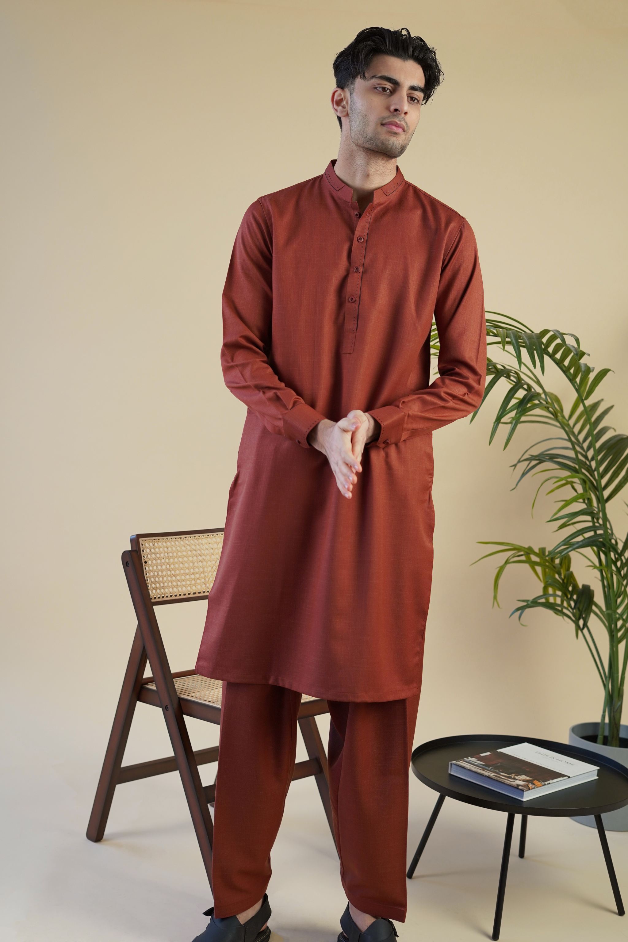 Pakistani male model wearing UD Fabrics red men's shalwar kameez with design on collar. Model poses with hands together in studio.