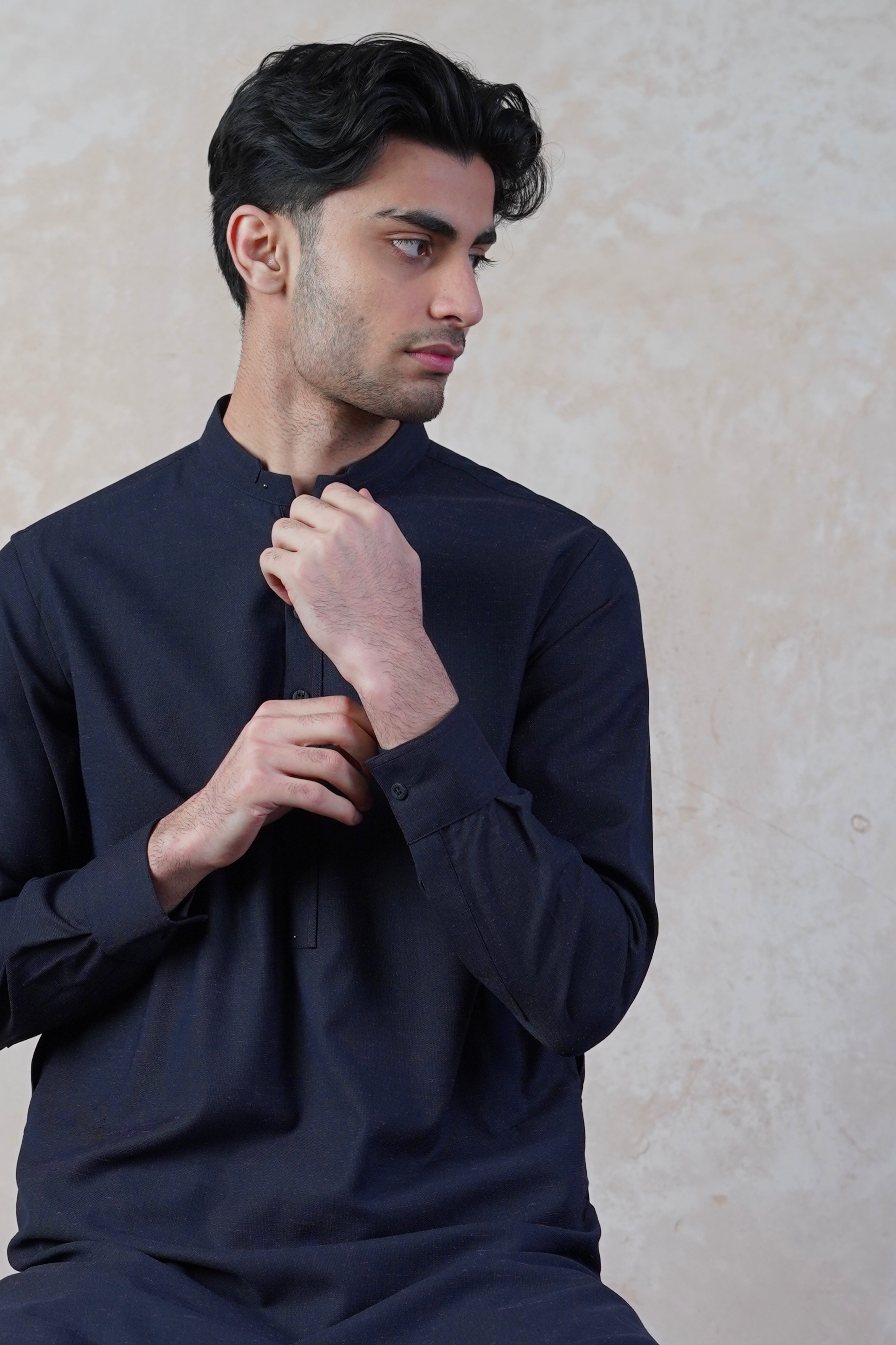 Pakistani male model standing wearing UD Fabrics men's navy blue shalwar kameez with purple undertones . Shalwar kameez with design on collar. Model poses looking away from camera. Men's pakistani clothing. Navy with purple undertones shalwar kameez. Design on collar of shalwar kameez.