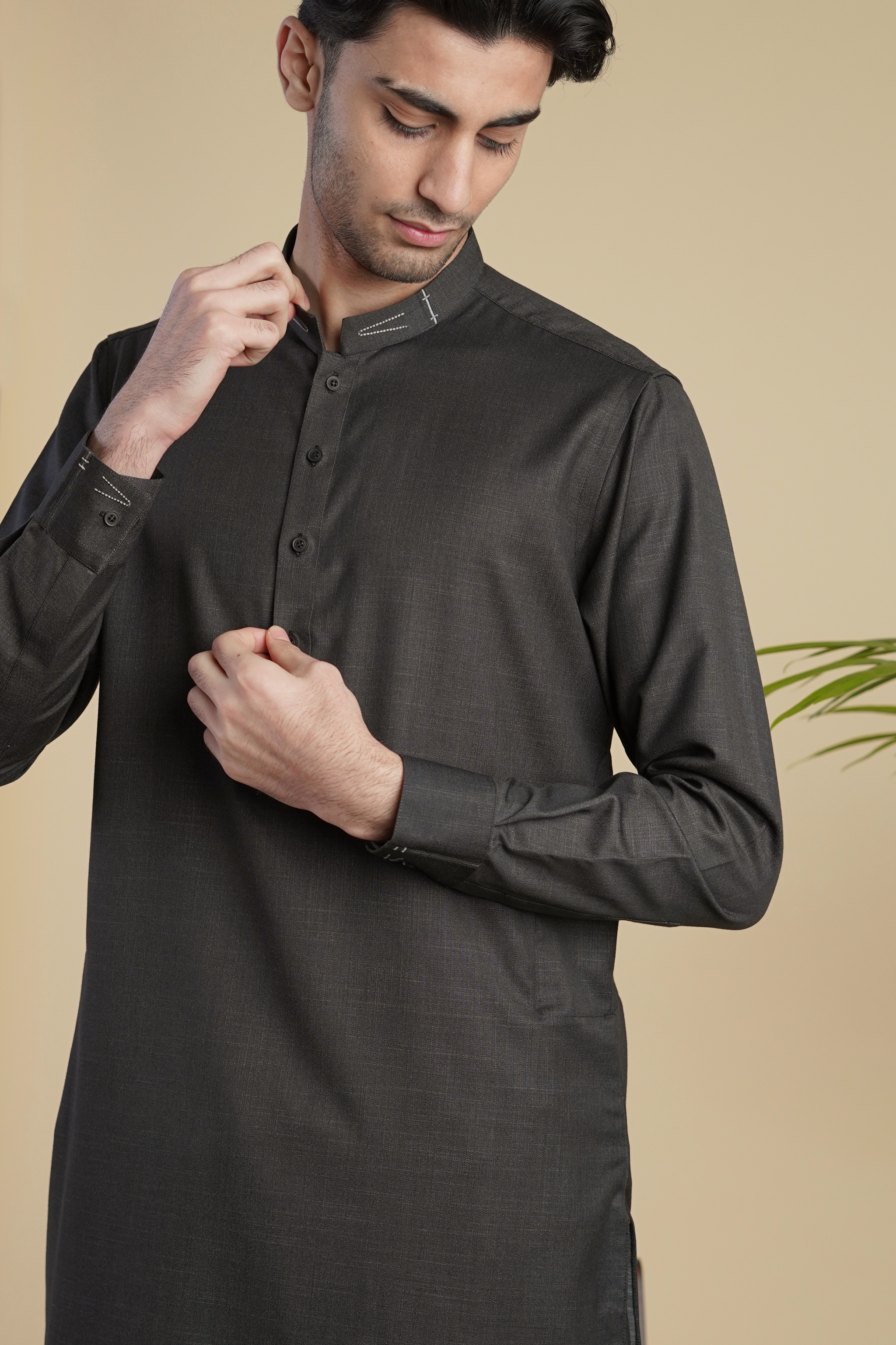 Pakistani male model standing wearing UD Fabrics men's black shalwar kameez. Shalwar kameez with design on collar. Model poses looking away from camera. Men's pakistani clothing. Black shalwar kameez