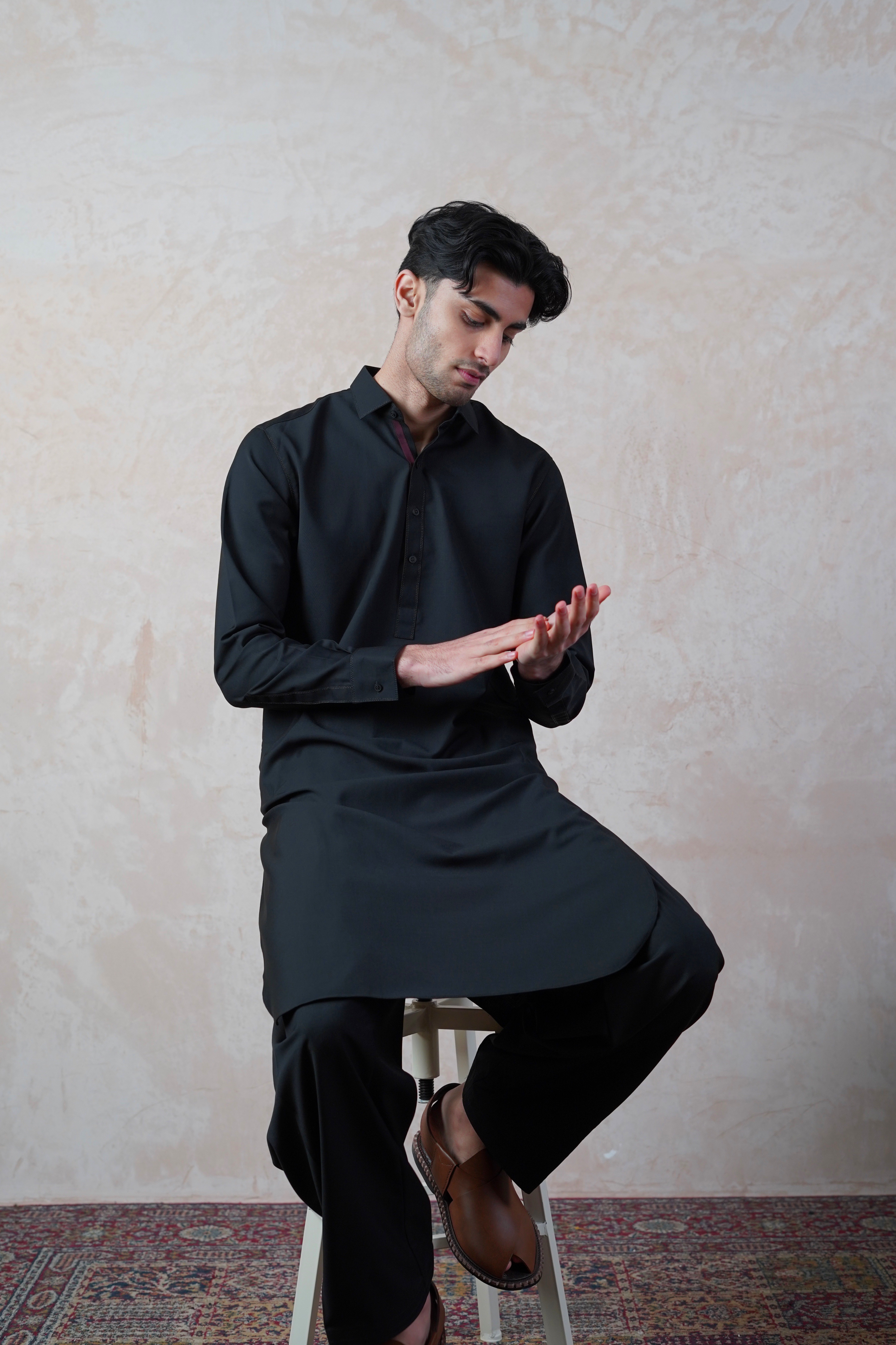 Pakistani male model sitting on stool wearing UD Fabrics men's black shalwar kameez. Shalwar kameez with full collar. Model poses looking towards hands. Men's pakistani clothing. Black shalwar kameez. Full collar shalwar kameez.