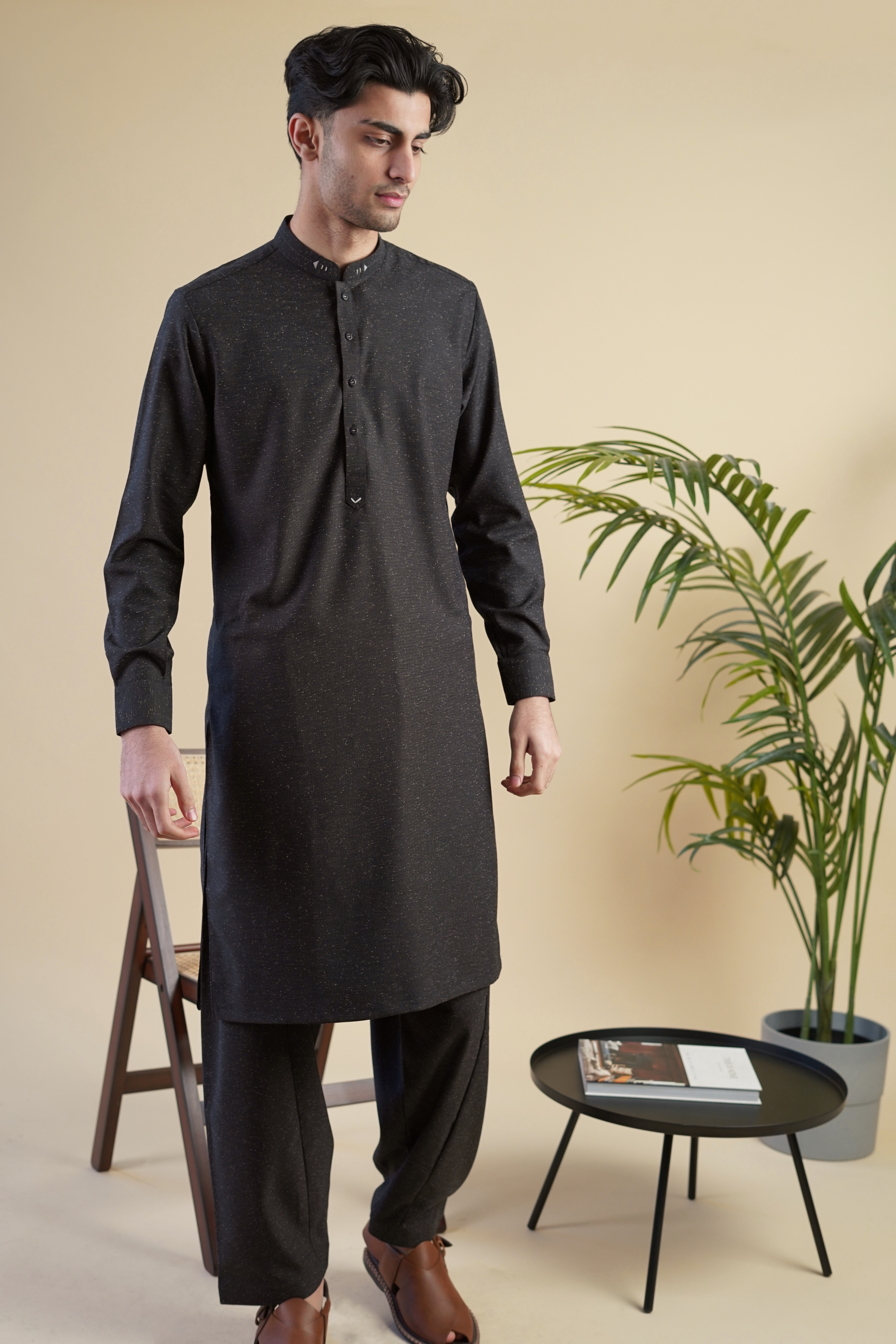 Pakistani male model standing wearing UD Fabrics men's black shalwar kameez. Shalwar kameez with design on collar. Model poses looking away from camera. Men's pakistani clothing. Black shalwar kameez with speckled design.