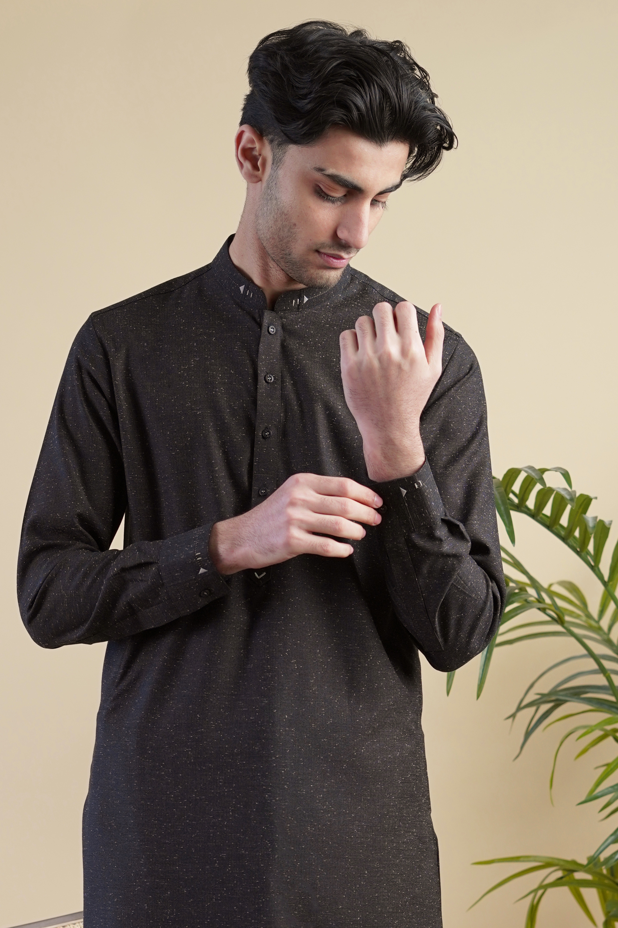 Detail image of Pakistani male model standing wearing UD Fabrics men's black shalwar kameez. Shalwar kameez with design on collar. Model poses looking away from camera. Men's pakistani clothing. Black shalwar kameez with speckled design. Design on shalwar kameez collar