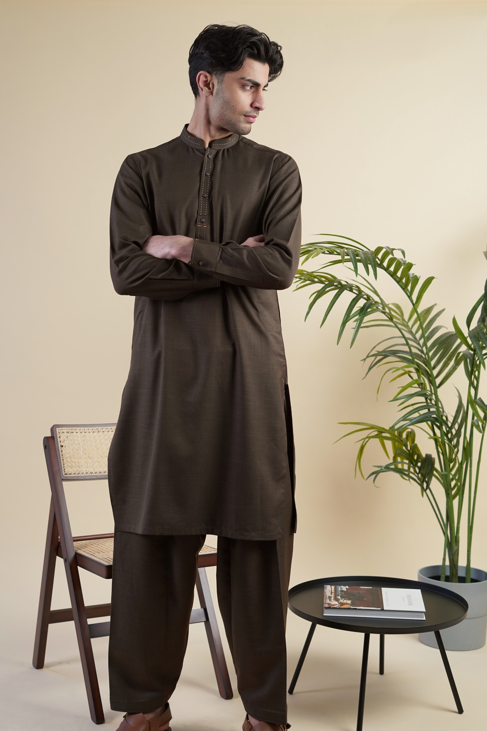 Pakistani male model standing wearing UD Fabrics men's brown shalwar kameez. Shalwar kameez with design on collar. Model poses looking away from camera. Men's pakistani clothing. Brown shalwar kameez.