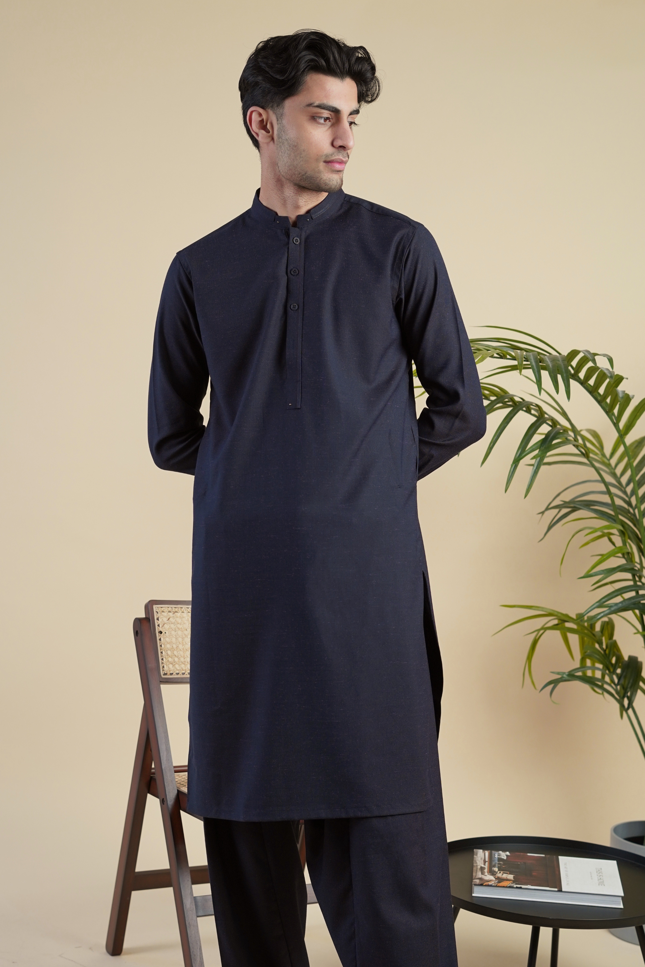 Pakistani male model standing wearing UD Fabrics men's navy blue shalwar kameez with purple undertones . Shalwar kameez with design on collar. Model poses looking away from camera. Men's pakistani clothing. Navy with purple undertones  shalwar kameez. Design on collar of shalwar kameez.