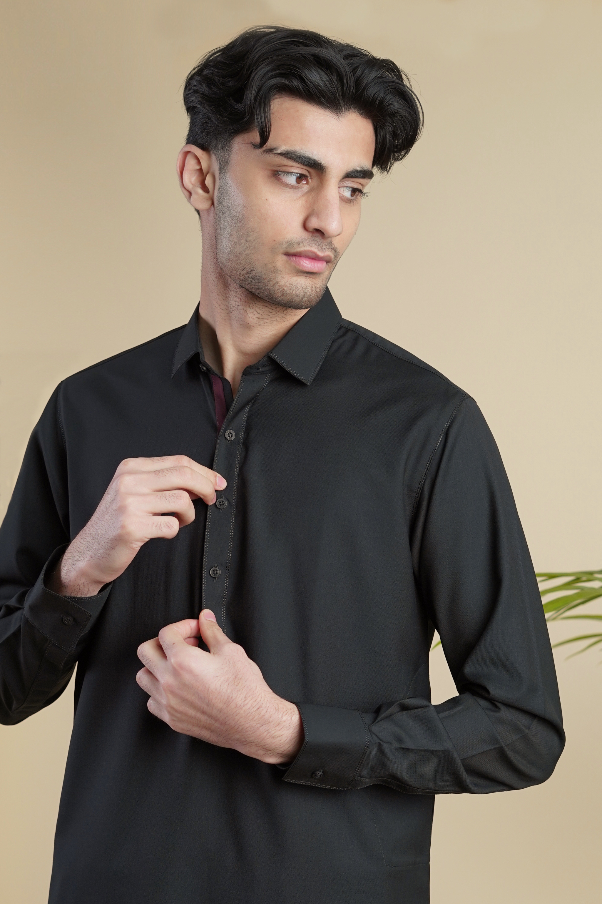 Pakistani male model standing wearing UD Fabrics men's black shalwar kameez. Shalwar kameez with full collar. Model poses looking away from camera. Men's pakistani clothing. Black shalwar kameez. Full collar shalwar kameez.