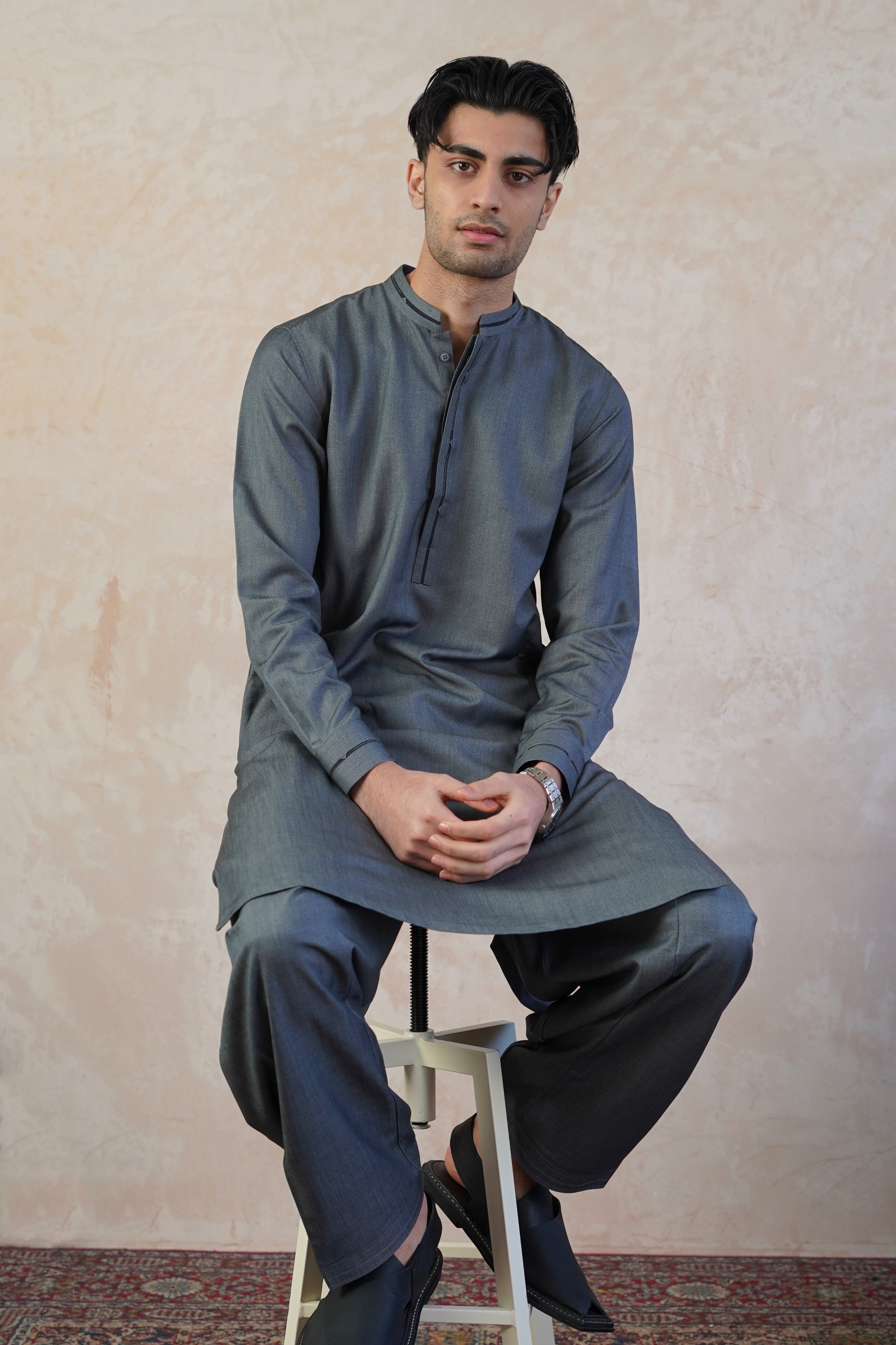 Pakistani male model wearing UD Fabrics shalwar kameez in grey with black band design on collar. Model sat of stool looking straight toward camera