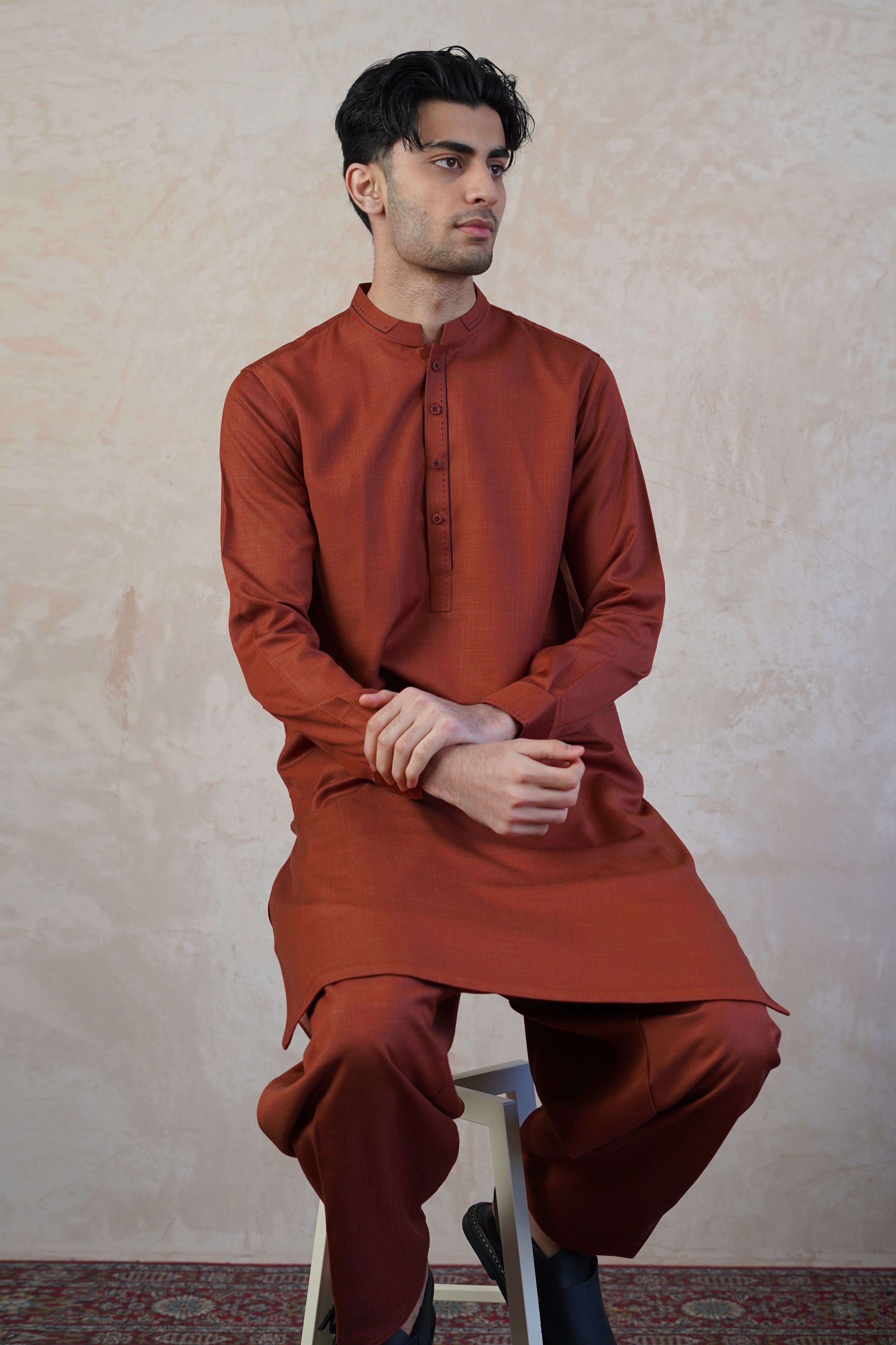 Pakistani male model wearing UD Fabrics men's red shalwar kameez with design on collar. Model sat of stool looking away from camera.