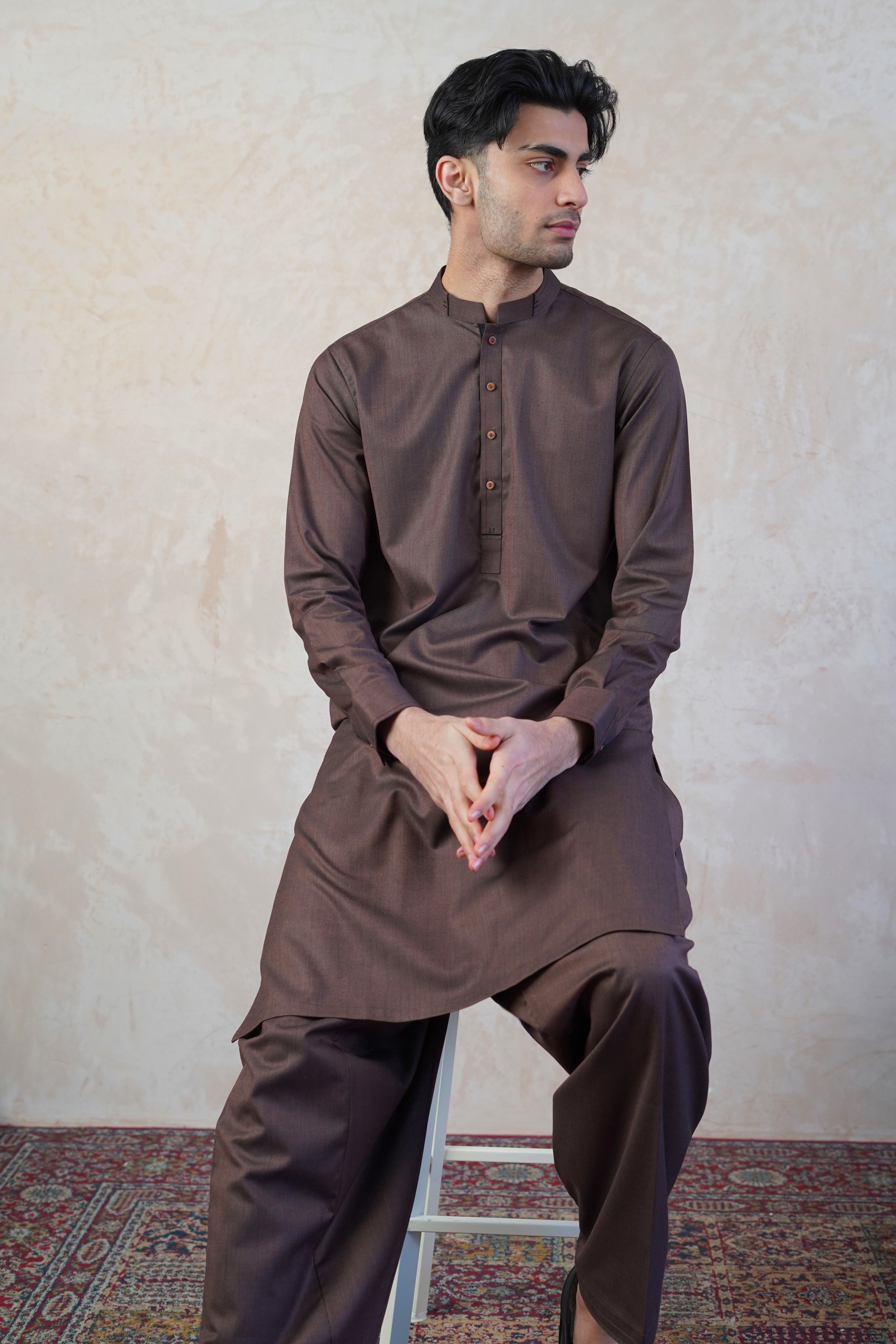 Pakistani male model wearing UD Fabrics men's brown shalwar kameez . Model sat on stool and poses looking away from camera