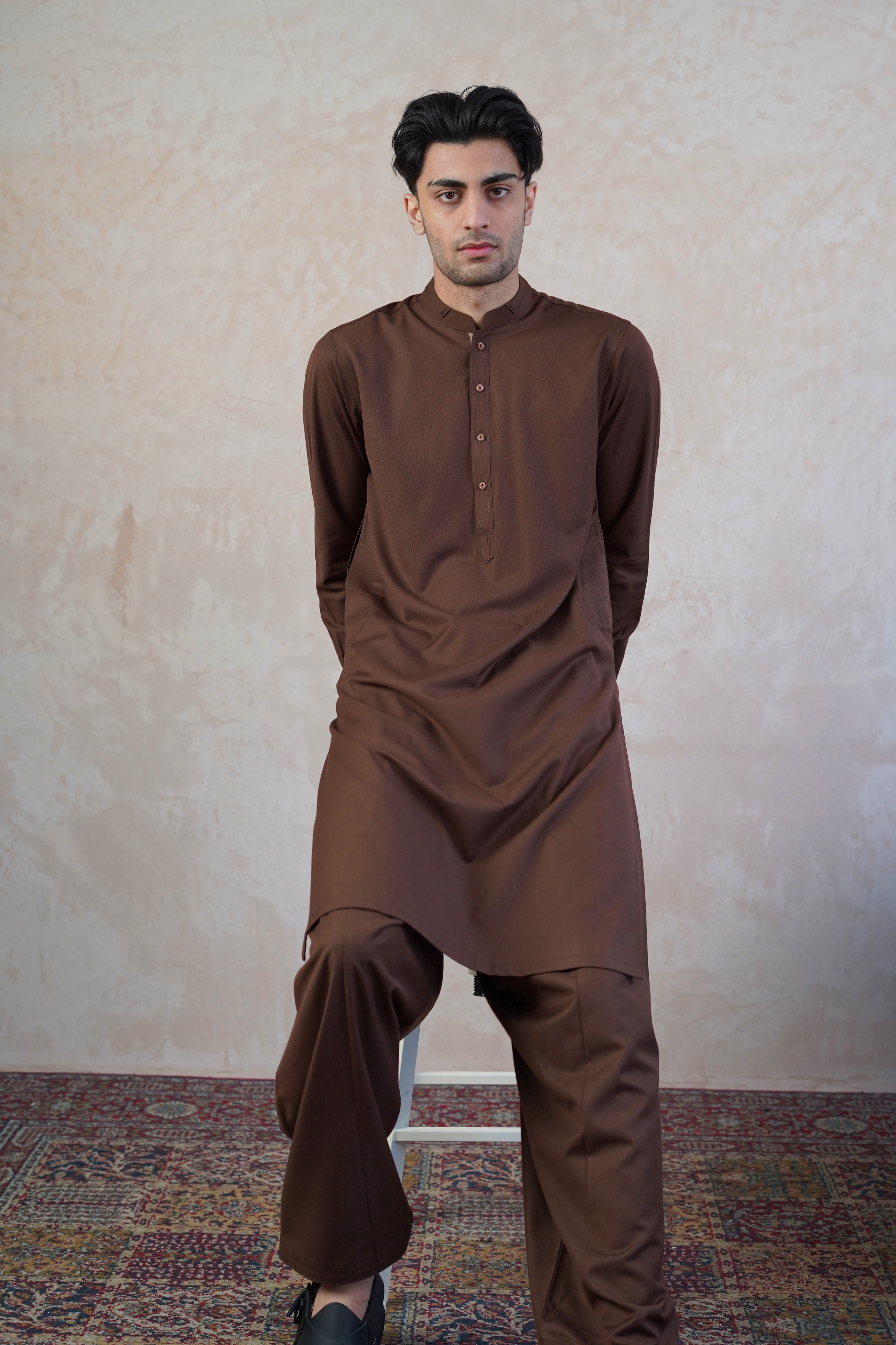 Pakistani male model sat on stool wearing UD Fabrics men's brown shalwar kameez. Shalwar kameez with design on collar. Model poses looking towards camera