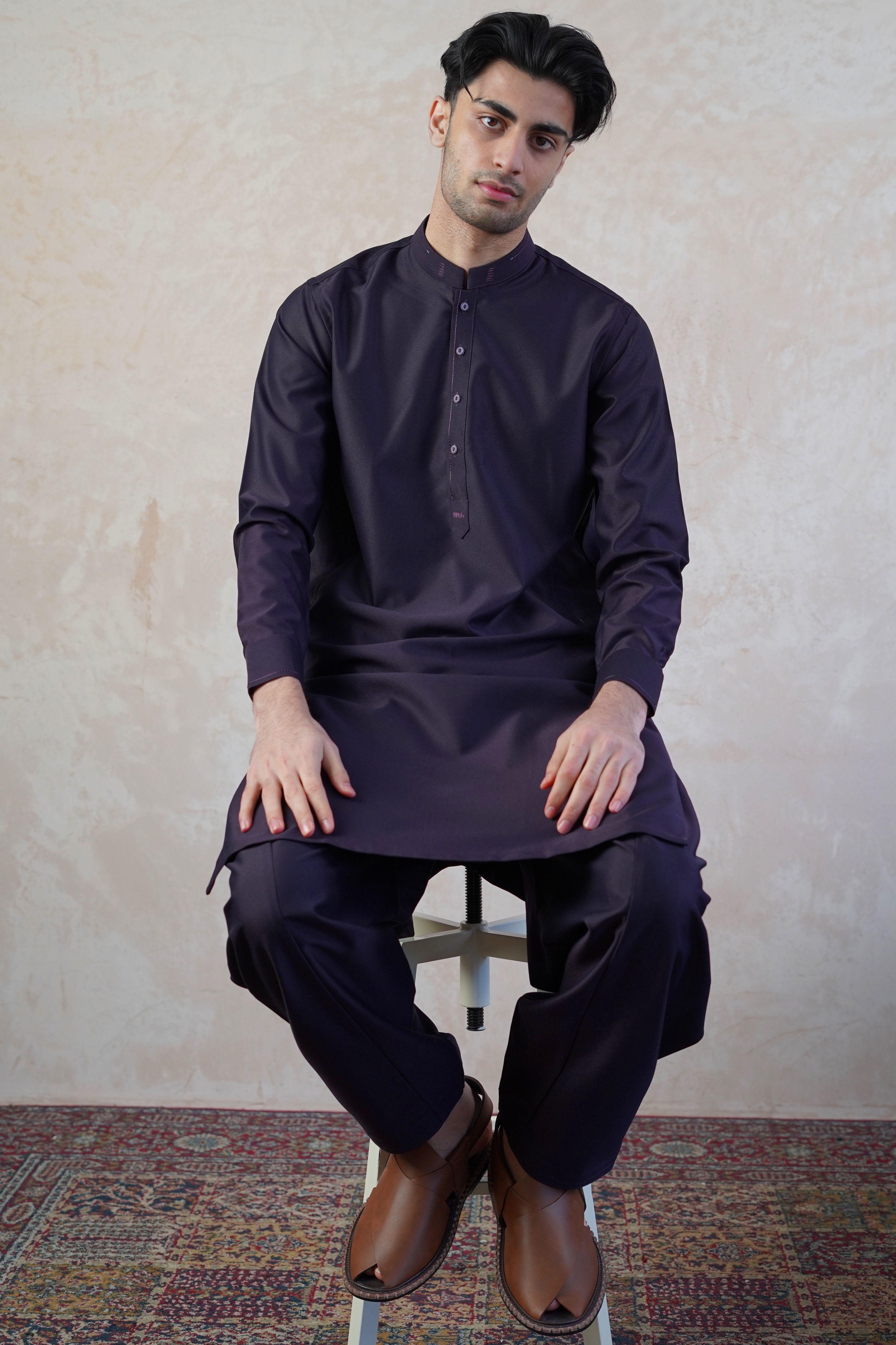 Pakistani male model sat on stool wearing UD Fabrics men's purple shalwar kameez. Shalwar kameez with design on collar. Model poses looking towards camera