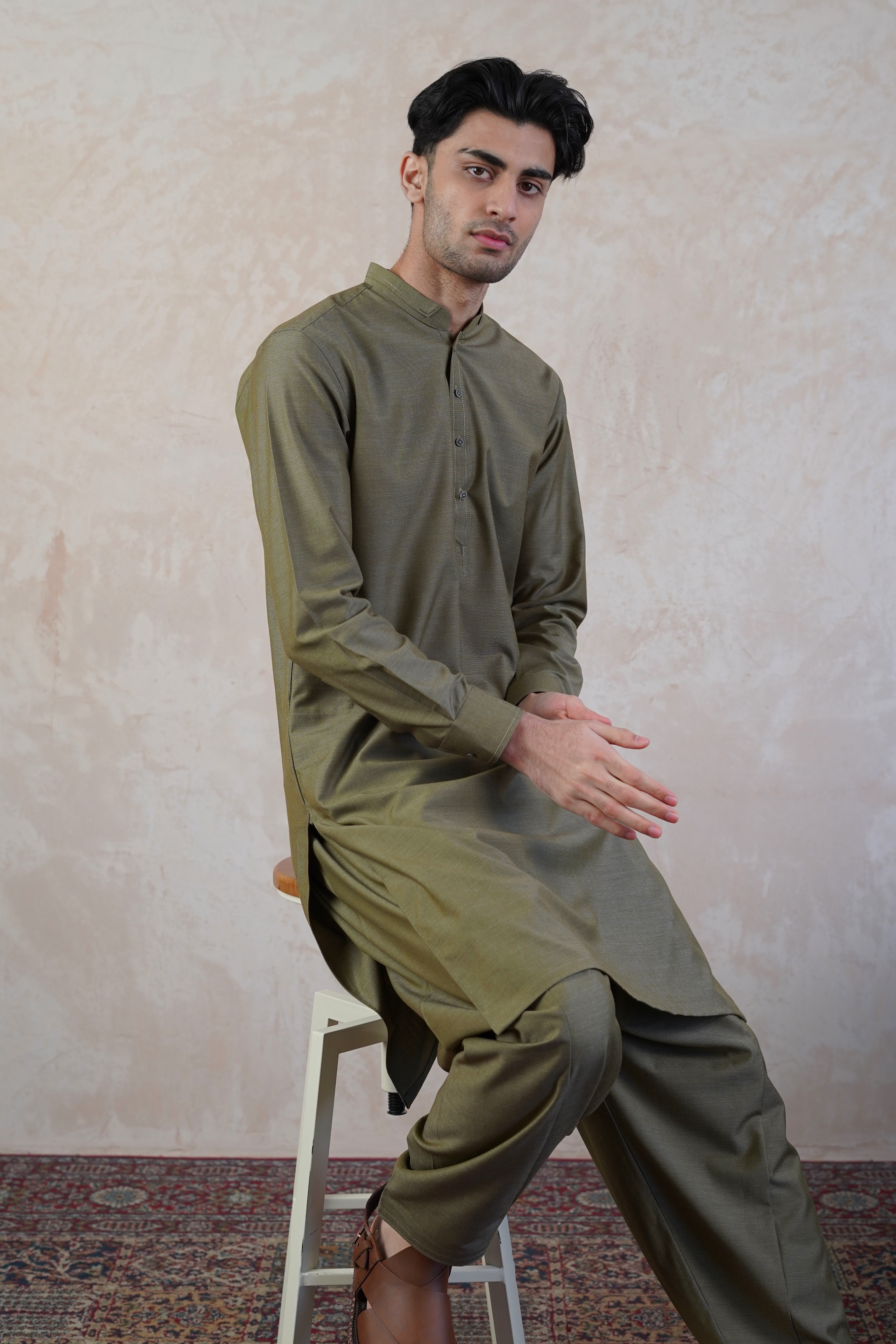 Pakistani male model sat on stool wearing UD Fabrics men's khaki green shalwar kameez. Shalwar kameez with design on collar. Model poses looking towards camera