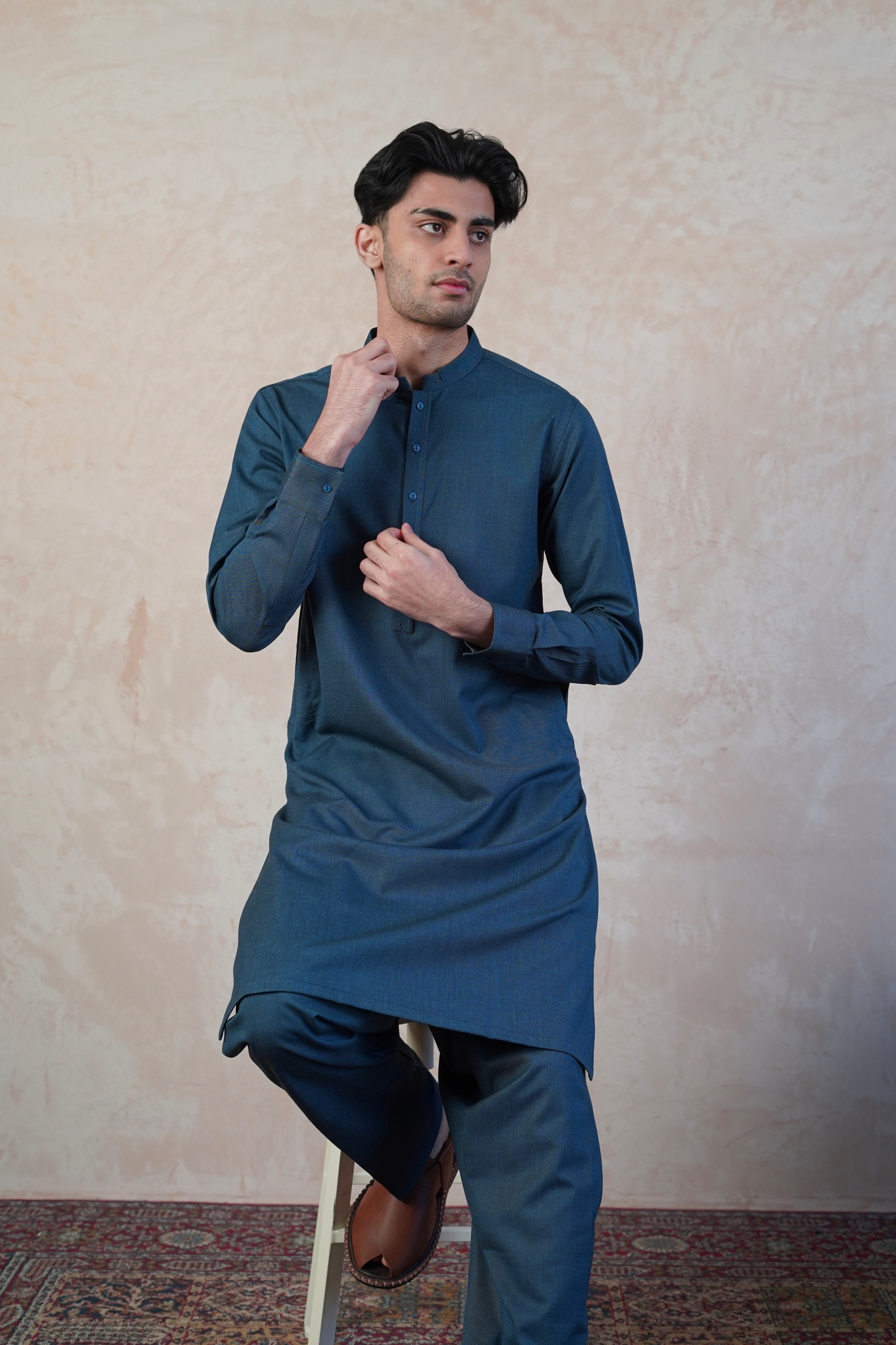 Pakistani male model sat on stool wearing UD Fabrics men's blue shalwar kameez. Shalwar kameez with design on collar. Model poses looking away from camera. Men's pakistani clothing. 