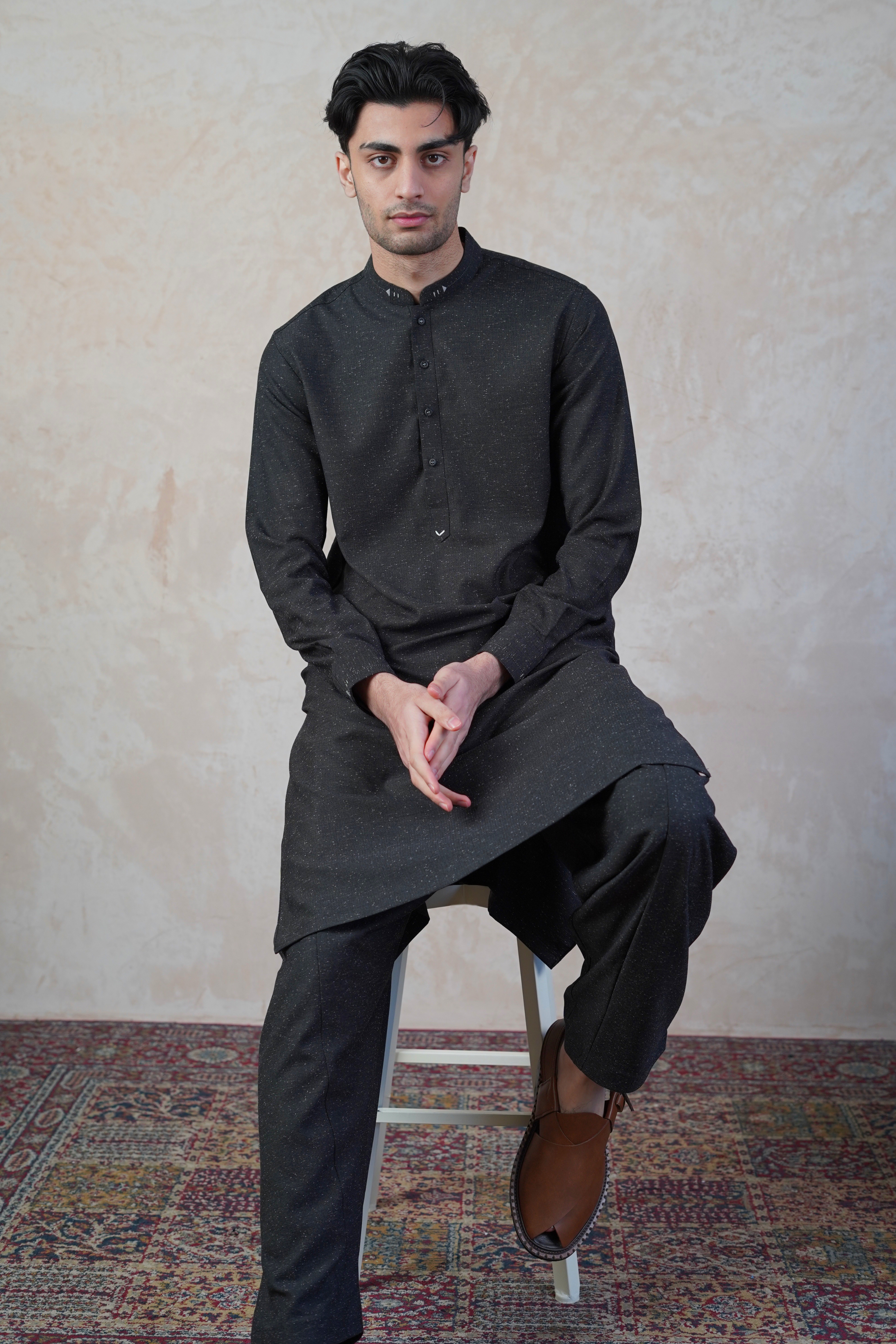 Pakistani male model sat on stool wearing UD Fabrics men's black shalwar kameez. Shalwar kameez with design on collar. Model poses looking directly toward camera. Men's pakistani clothing. Black shalwar kameez with speckled design.