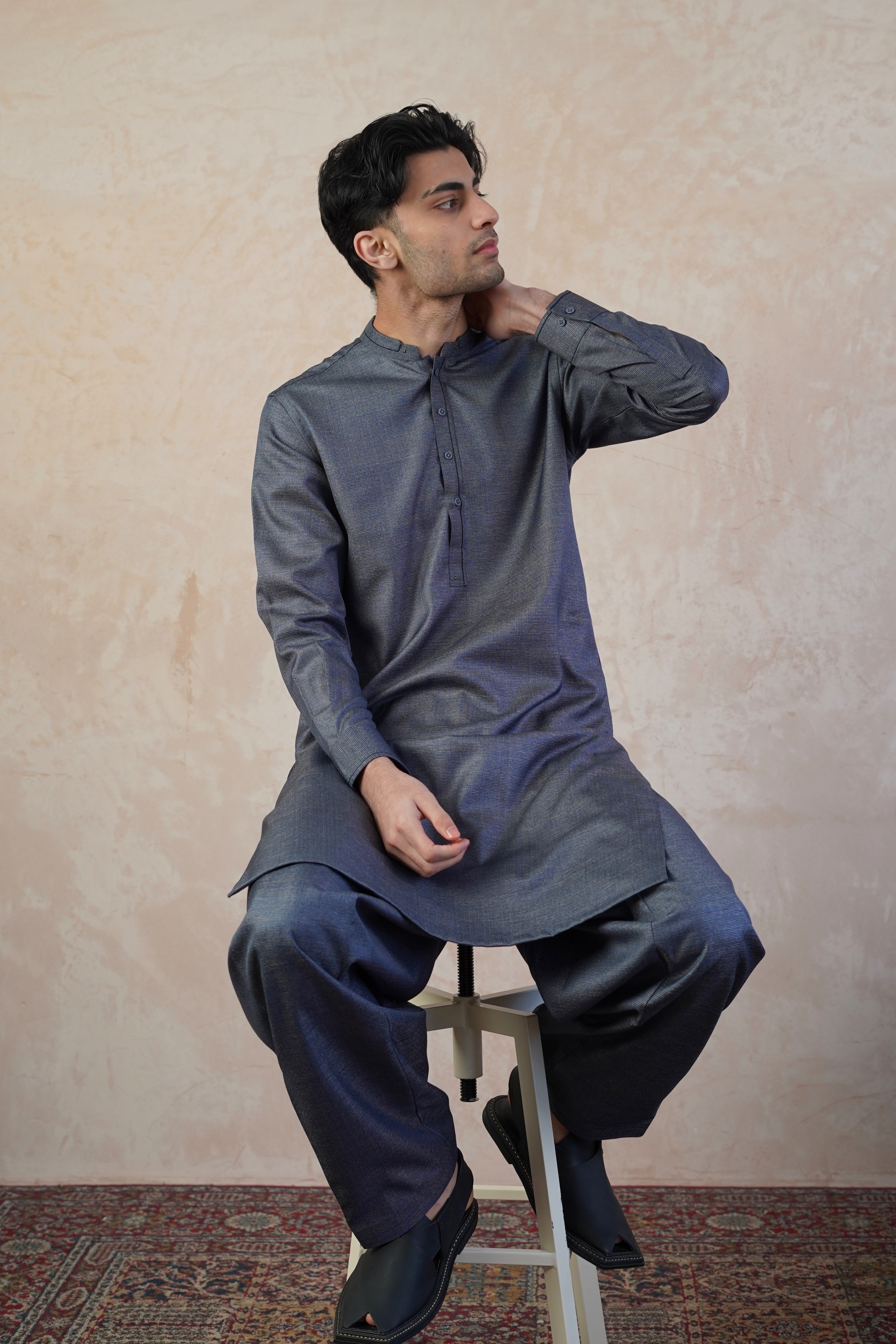 Pakistani male model sat on stool wearing UD Fabrics men's grey shalwar kameez. Shalwar kameez with design on collar. Model poses looking away from camera. Men's pakistani clothing